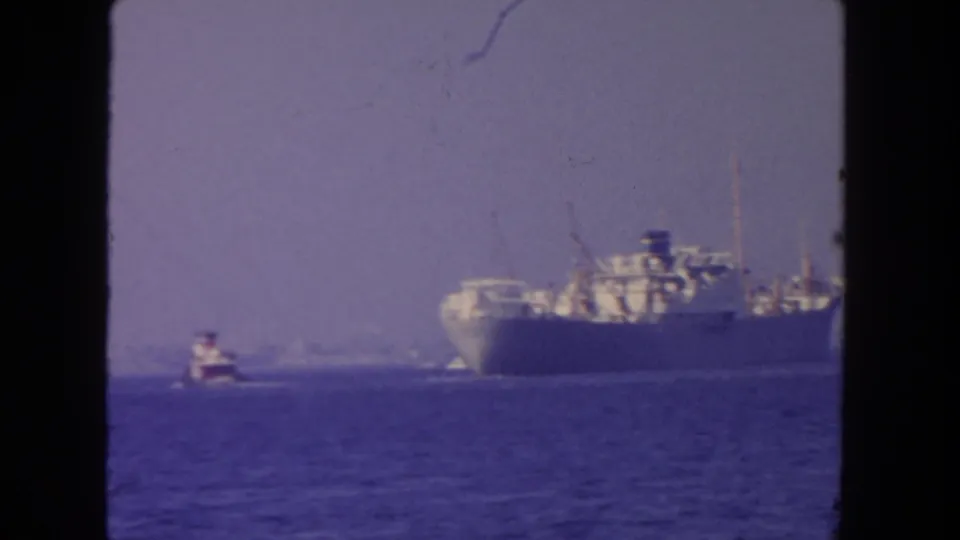 1962 San Diego 8mm frame showing a side view of a cargo ship with deck structures and a tugboat on calm water.