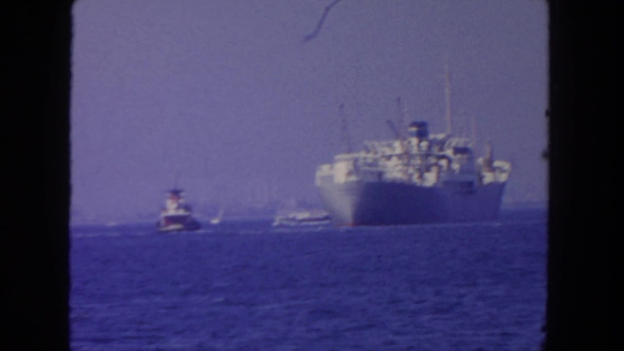 1962 San Diego home-movie frame showing a distant large ship and small tugboat on blue water beneath a hazy sky.