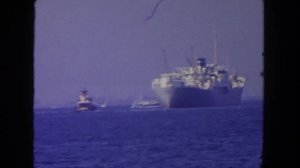 1962 San Diego home-movie frame showing a distant large ship and small tugboat on blue water beneath a hazy sky.