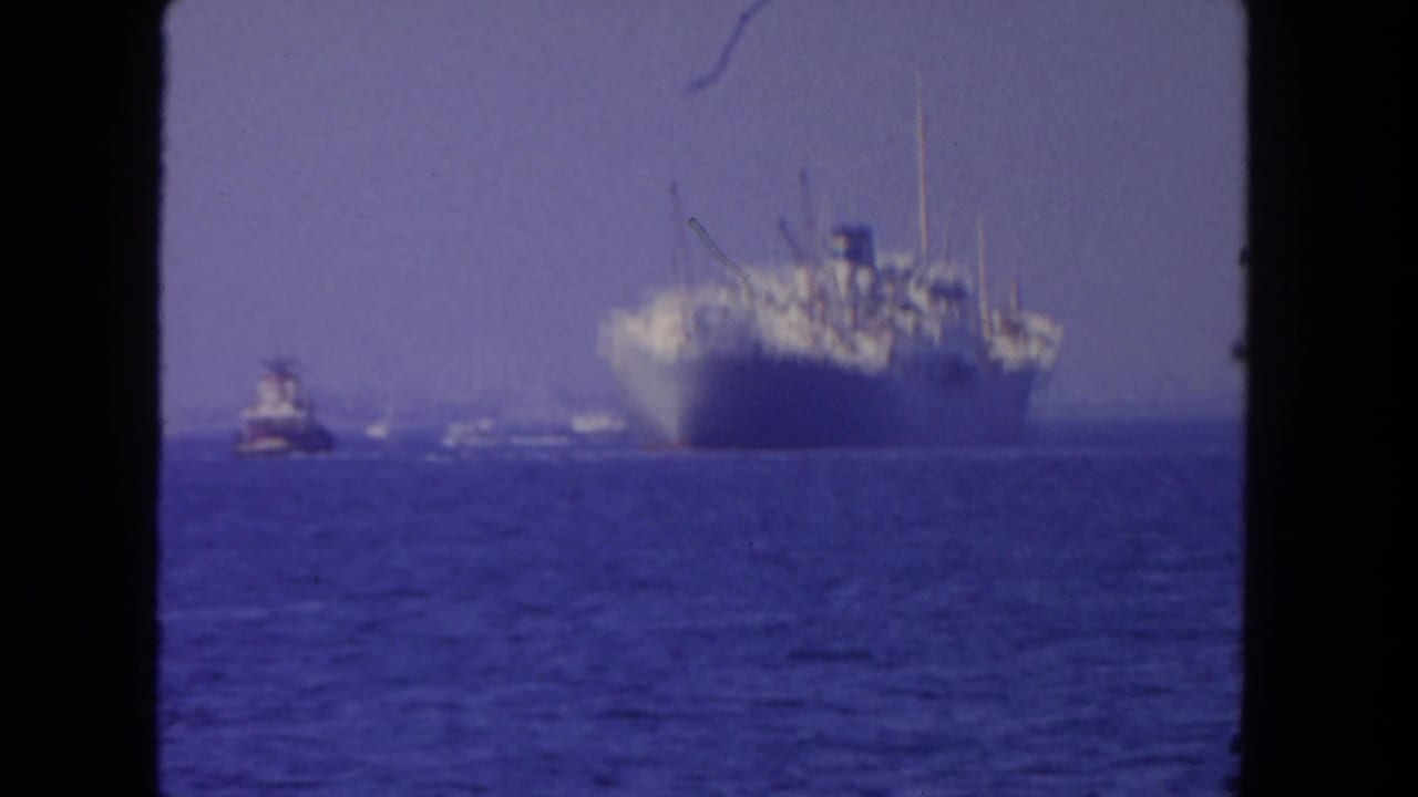 1962 San Diego 8mm frame with a large gray ship on open water, accompanied by a tugboat and distant small craft.