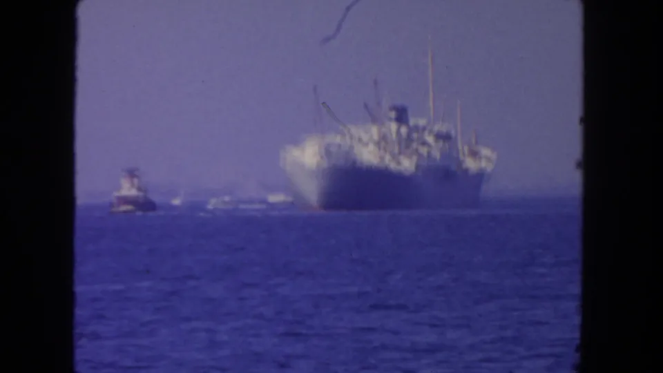 1962 San Diego 8mm frame with a large gray ship on open water, accompanied by a tugboat and distant small craft.