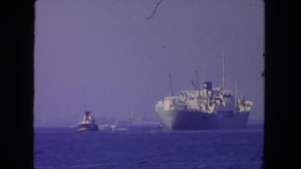 1962 San Diego 8mm frame placing a large vessel near the right edge, with a tugboat on blue ocean in haze.