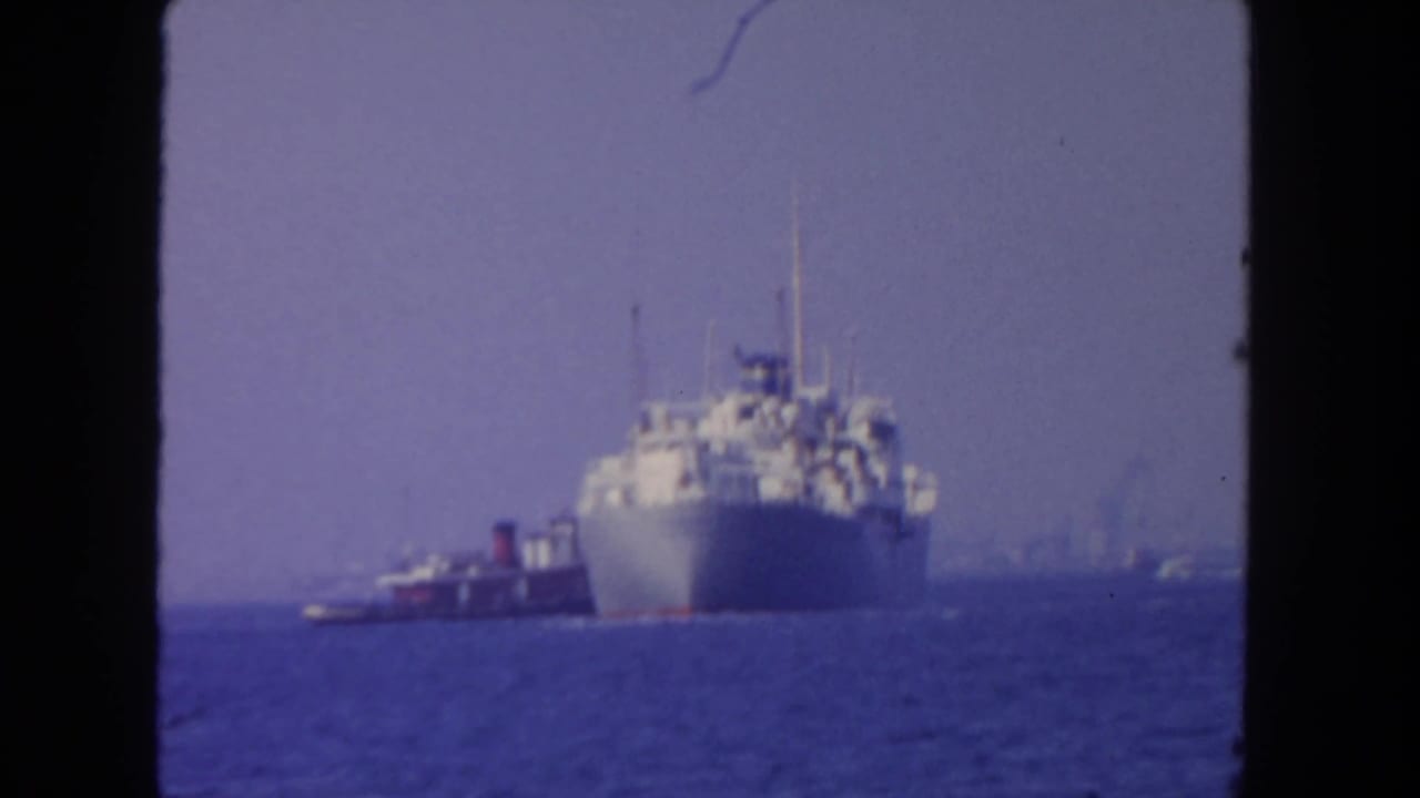 Passenger ship and tugboat seen at a distance across blue water with haze in the background.