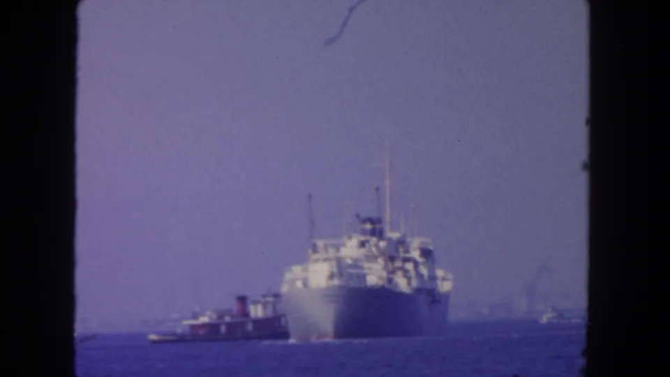 1962 home-movie frame of a large passenger ship offshore, with tugboat support and muted blue-purple 8mm film color.