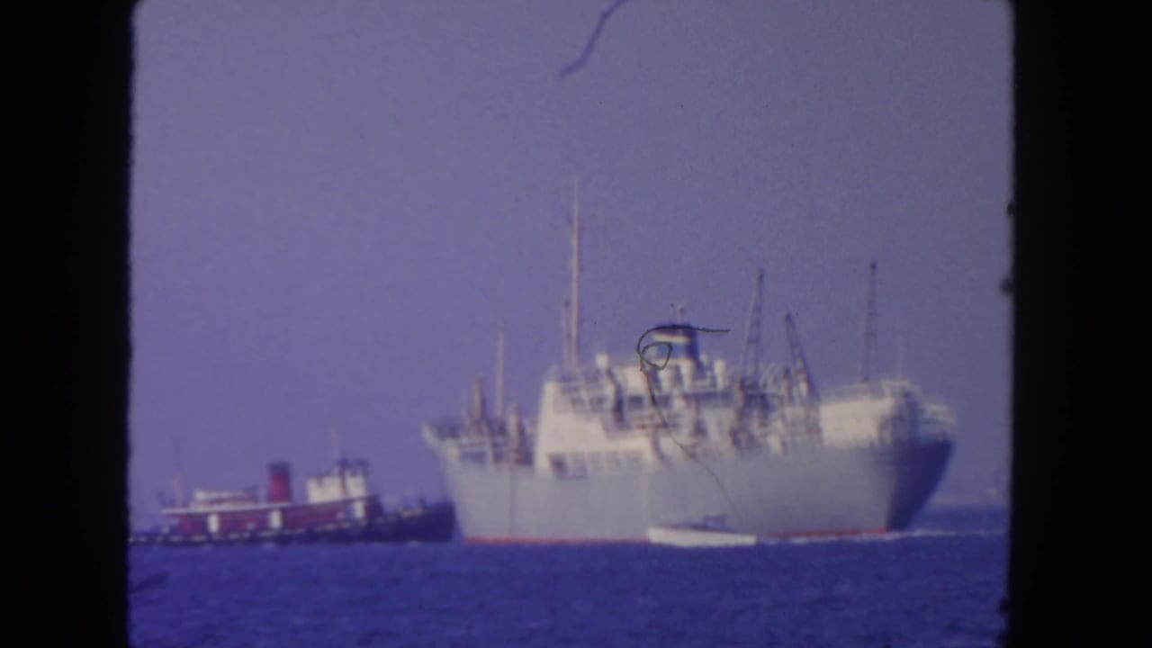 1962 San Diego 8mm frame showing a large cargo ship with a tugboat nearby on blue ocean water.
