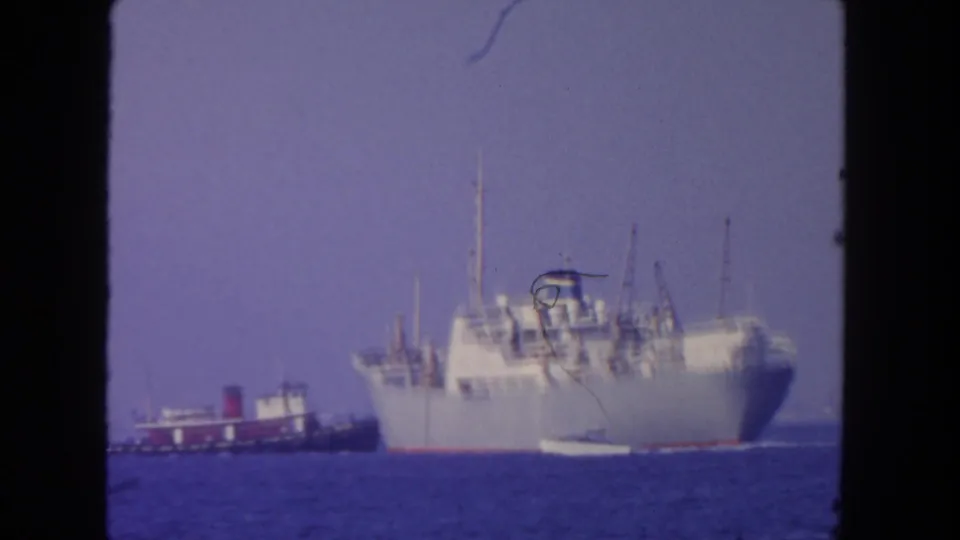 1962 San Diego 8mm frame showing a large cargo ship with a tugboat nearby on blue ocean water.