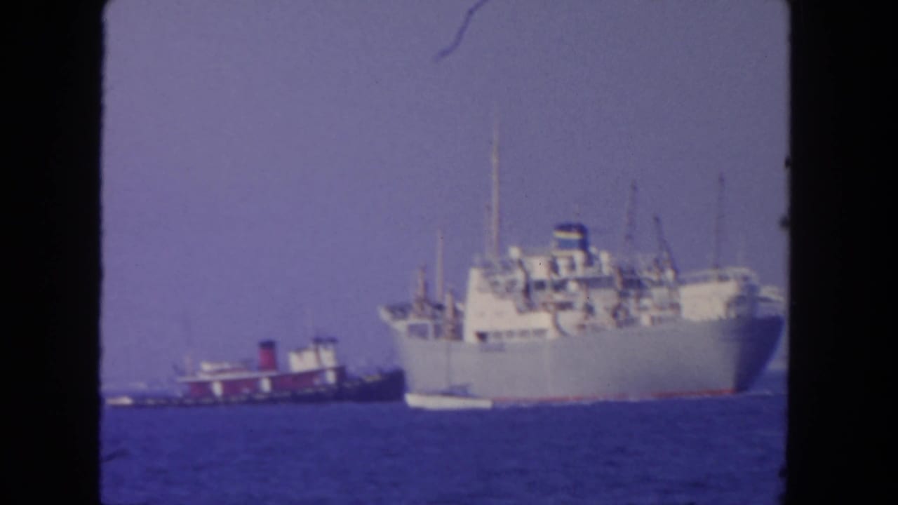 1962 San Diego home-movie frame of a large freighter and tugboat moving across blue water.