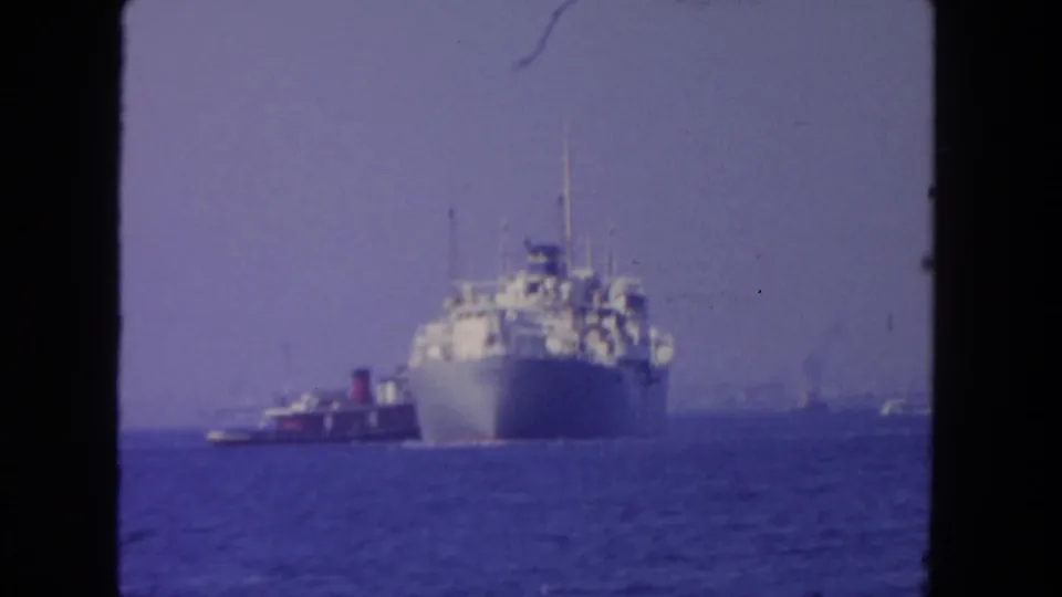 1962 San Diego 8mm frame showing the bow of a cargo ship with a tugboat beside it on hazy blue water.