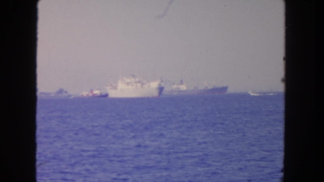 1962 San Diego-area 8mm view of distant naval ships floating offshore under a hazy sky, framed by dark edges.