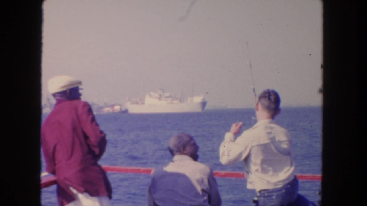 1962 San Diego 8mm frame showing three men aboard a boat, one holding a fishing rod, with large ships on the horizon.