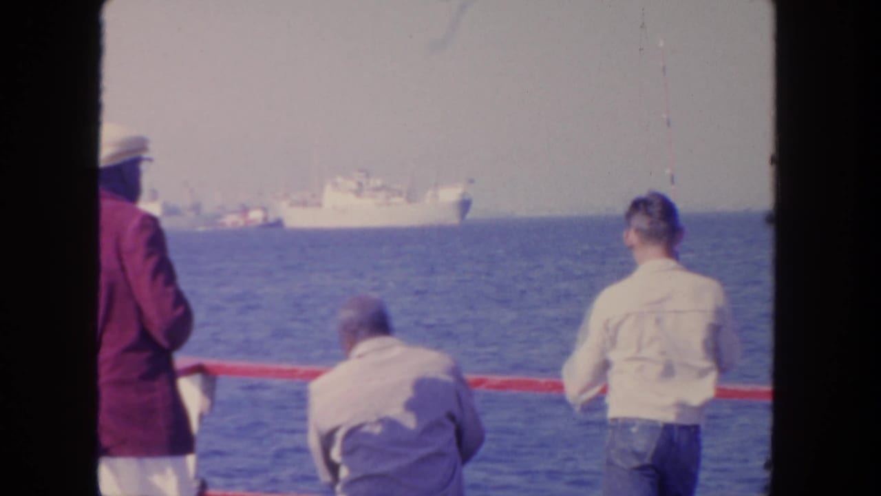 1962 San Diego home-movie frame with men at a boat rail watching distant white ships over calm water.