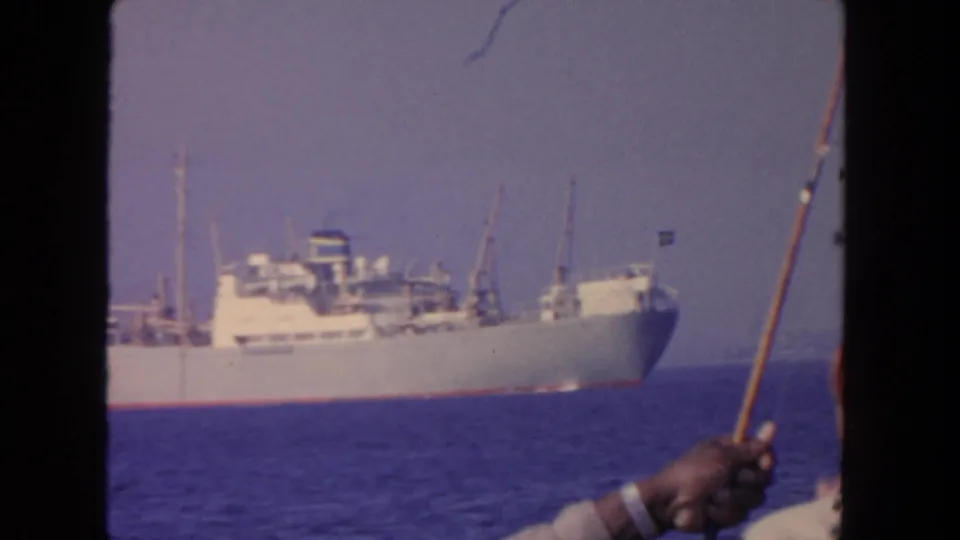 1962 San Diego home-movie frame showing a cargo ship on blue water, seen past a raised fishing rod and partial foreground figures.