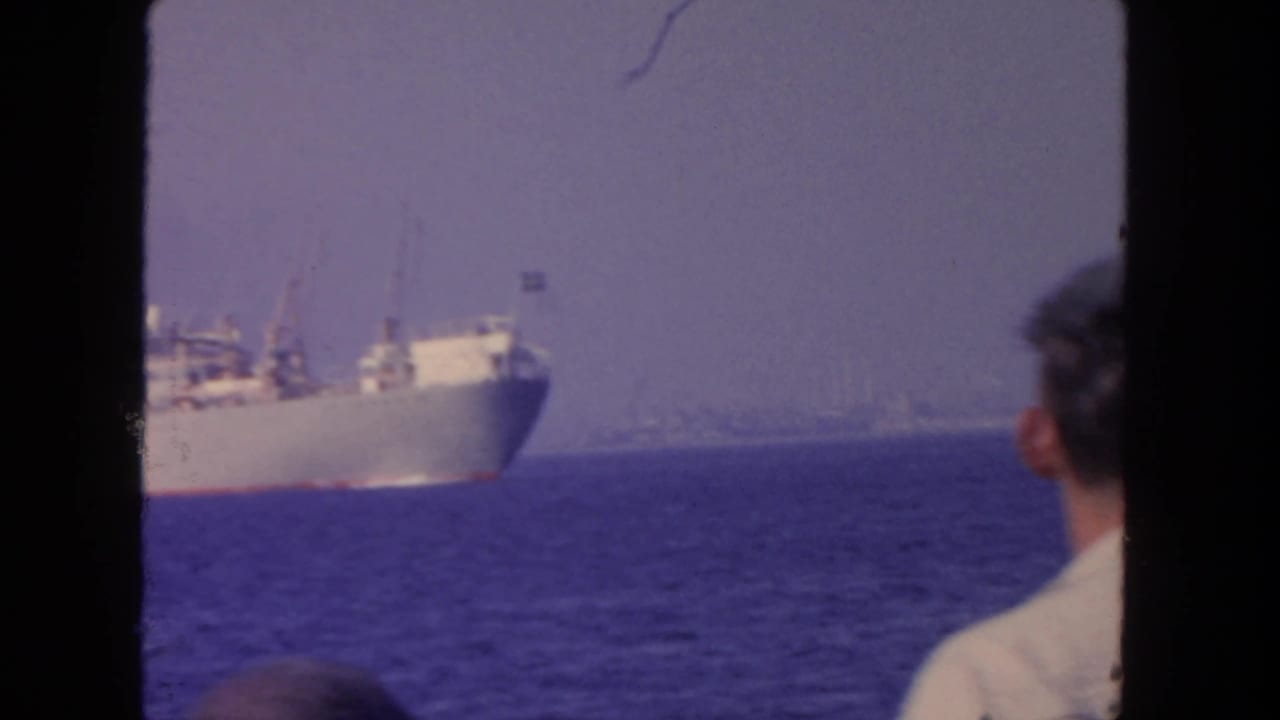 Cargo ship on the water with a seated or standing man seen from behind at right.