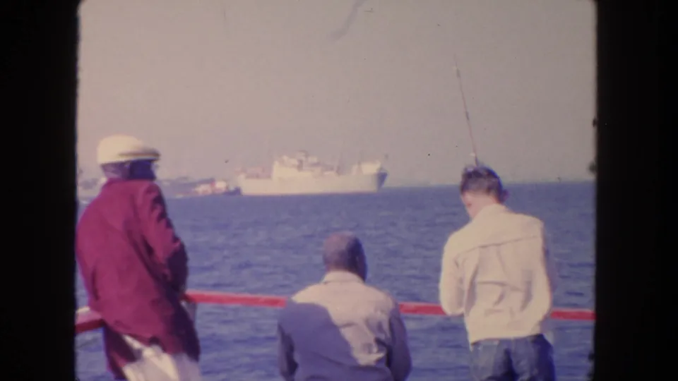 1962 San Diego home-movie frame showing men by a red railing, one with a fishing rod, and distant ships on the water.