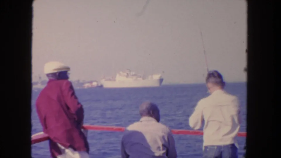 1962 San Diego frame of men seen from behind at a red railing, with fishing rods and a cargo ship offshore.