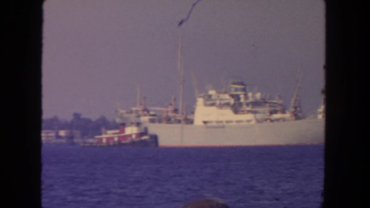 1962 San Diego harbor frame showing a white cargo ship approached by a red-and-white tugboat under a hazy sky.