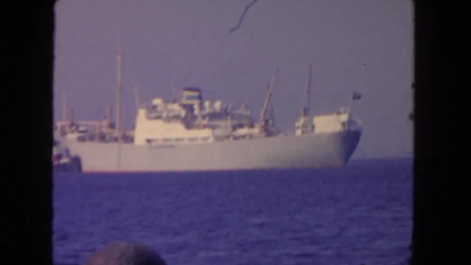 1962 San Diego harbor frame showing the cargo ship farther across the water, with tugboat at left and incidental foreground head.