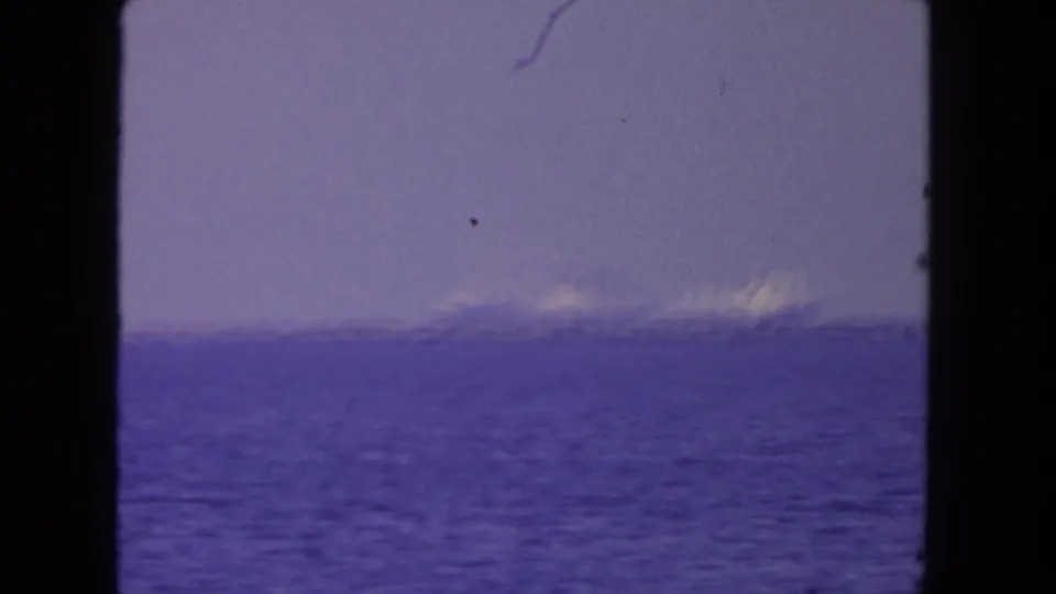 1962 San Diego 8mm frame showing distant cargo vessels across blue water under a hazy purple sky.