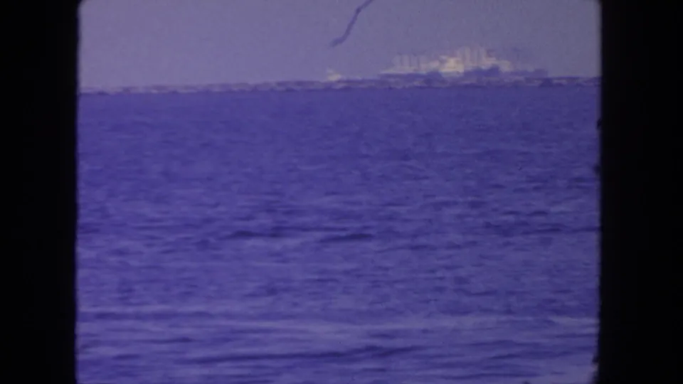 1962 8mm coastal frame with a large passenger ship far offshore, calm water in front, and a hazy sky behind it.