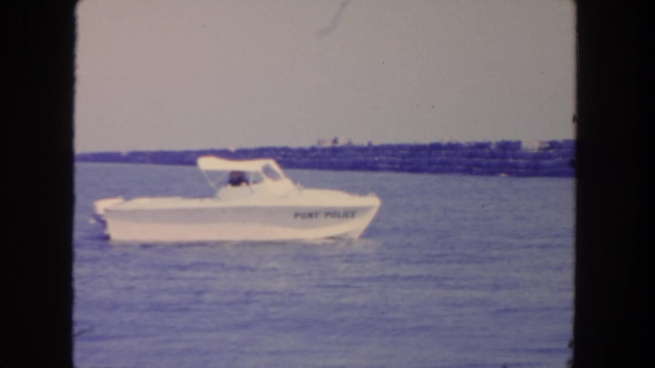 White port police boat moves across San Diego harbor water near a low breakwater in 1962 home-movie footage.