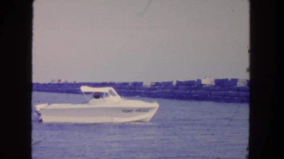 White port police boat cruising on calm San Diego harbor water with breakwater rocks behind it, filmed on 8mm in 1962.
