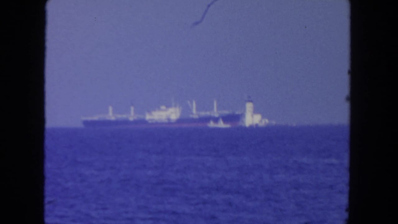 1962 San Diego 8mm frame showing a distant cargo ship and smaller vessel on open water beneath a hazy blue sky.