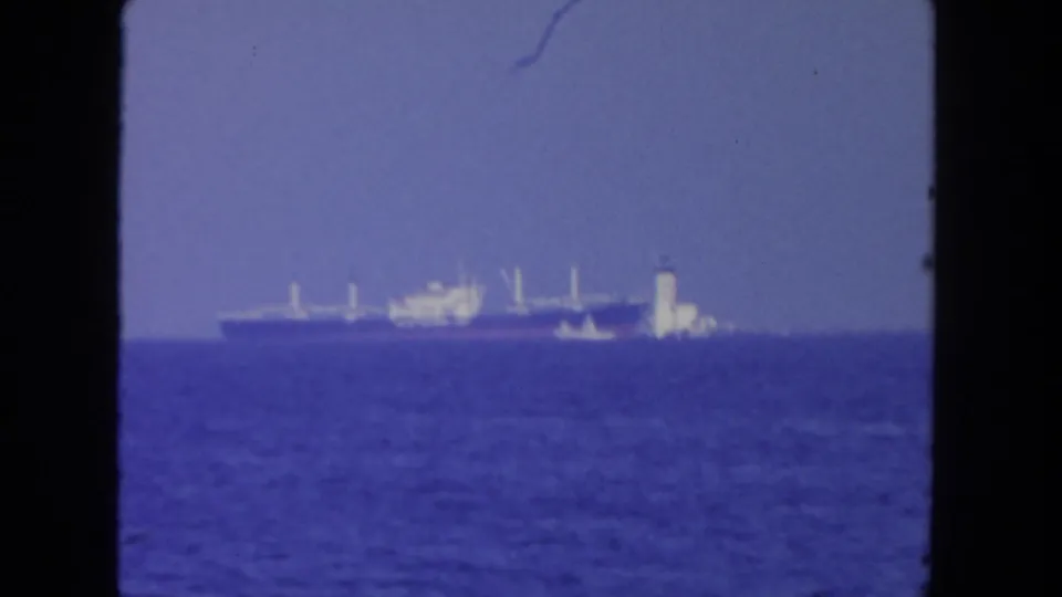 1962 San Diego 8mm frame showing a distant cargo ship and smaller vessel on open water beneath a hazy blue sky.