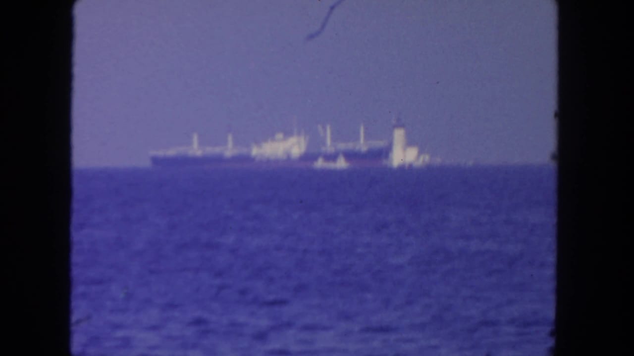 Distant 1962 San Diego ocean view with a cargo ship and nearby smaller vessel seen through 8mm film framing.