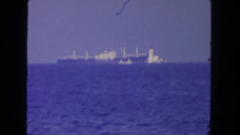 Distant 1962 San Diego ocean view with a cargo ship and nearby smaller vessel seen through 8mm film framing.