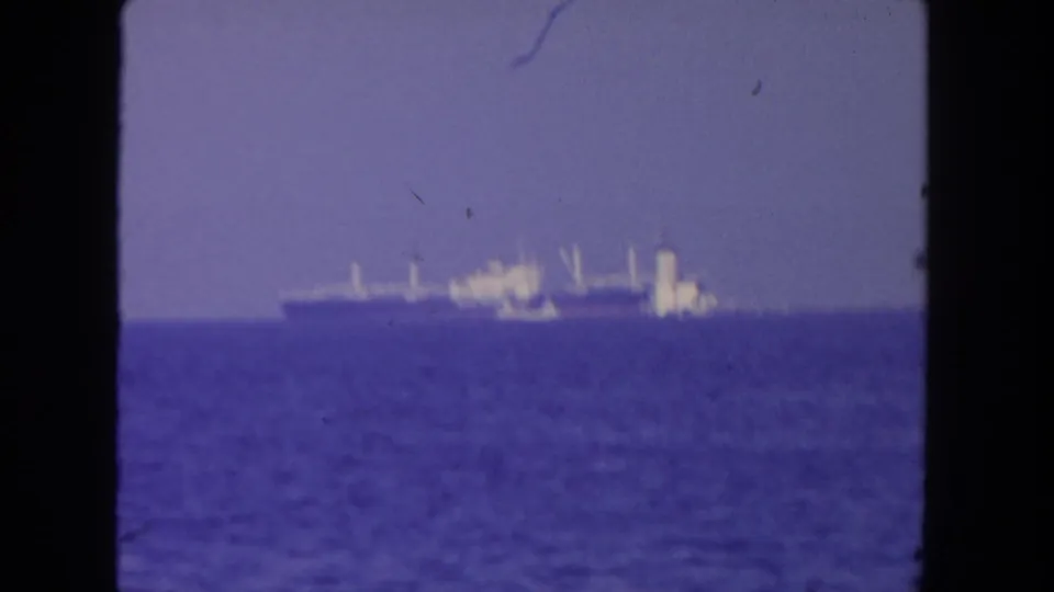Grainy 8mm frame from the San Diego clip showing a distant cargo ship, smaller vessel, ocean, and hazy sky.