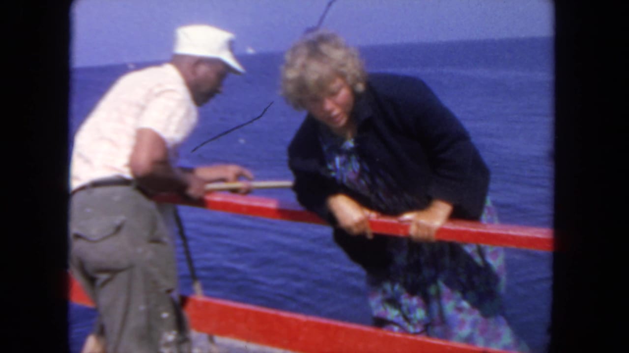 In 1962 San Diego home-movie footage, a woman in a blue patterned dress climbs over a red boat rail as a man in a white cap helps beside the ocean.