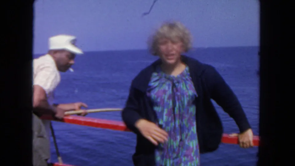 The woman in a blue patterned dress faces the camera after boarding, with the man partly visible beside the red rail and ocean horizon.