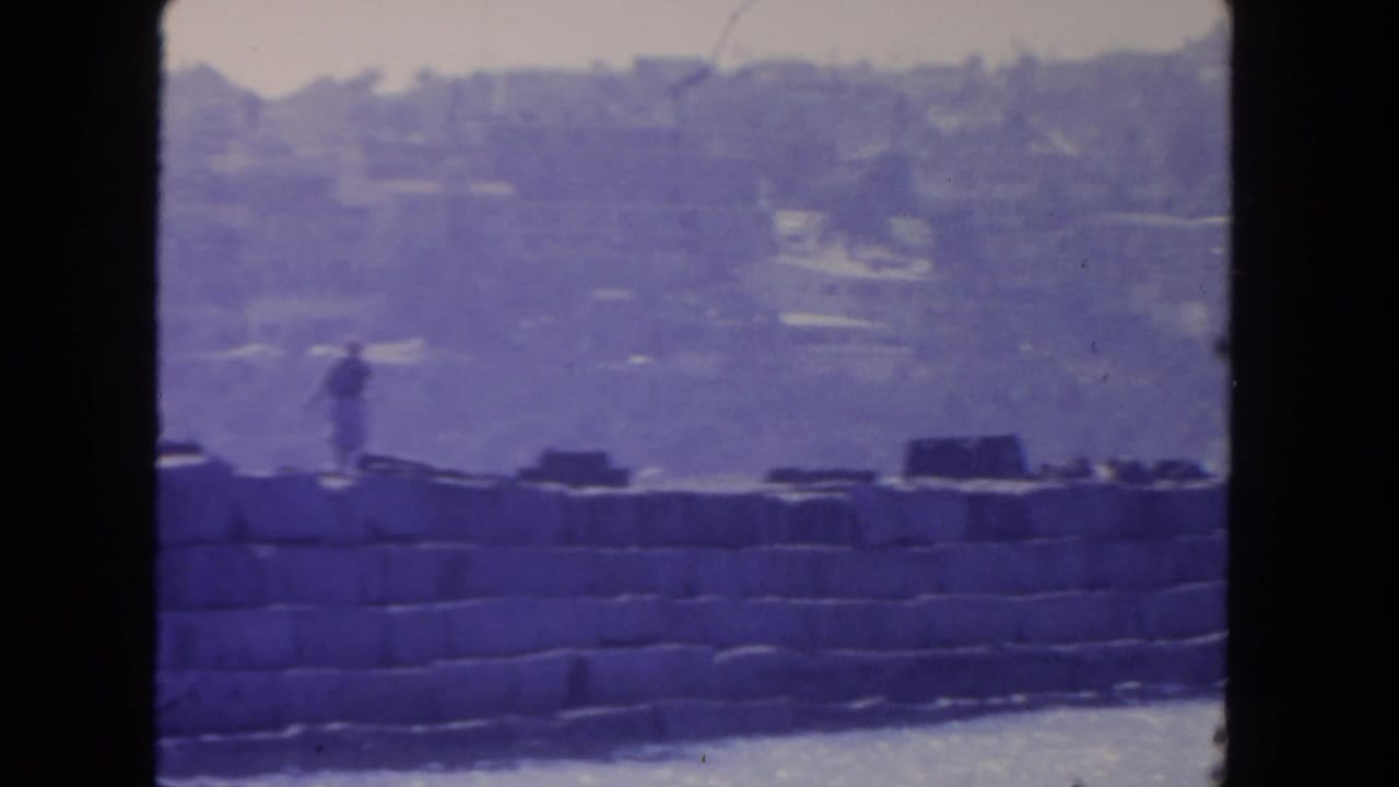 Distant figure walking along a stone seawall beside water under a hazy shoreline.