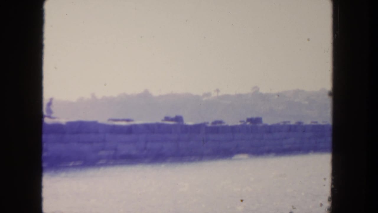 1962 San Diego 8mm frame showing a small distant figure running left to right along a stone seawall beside water.