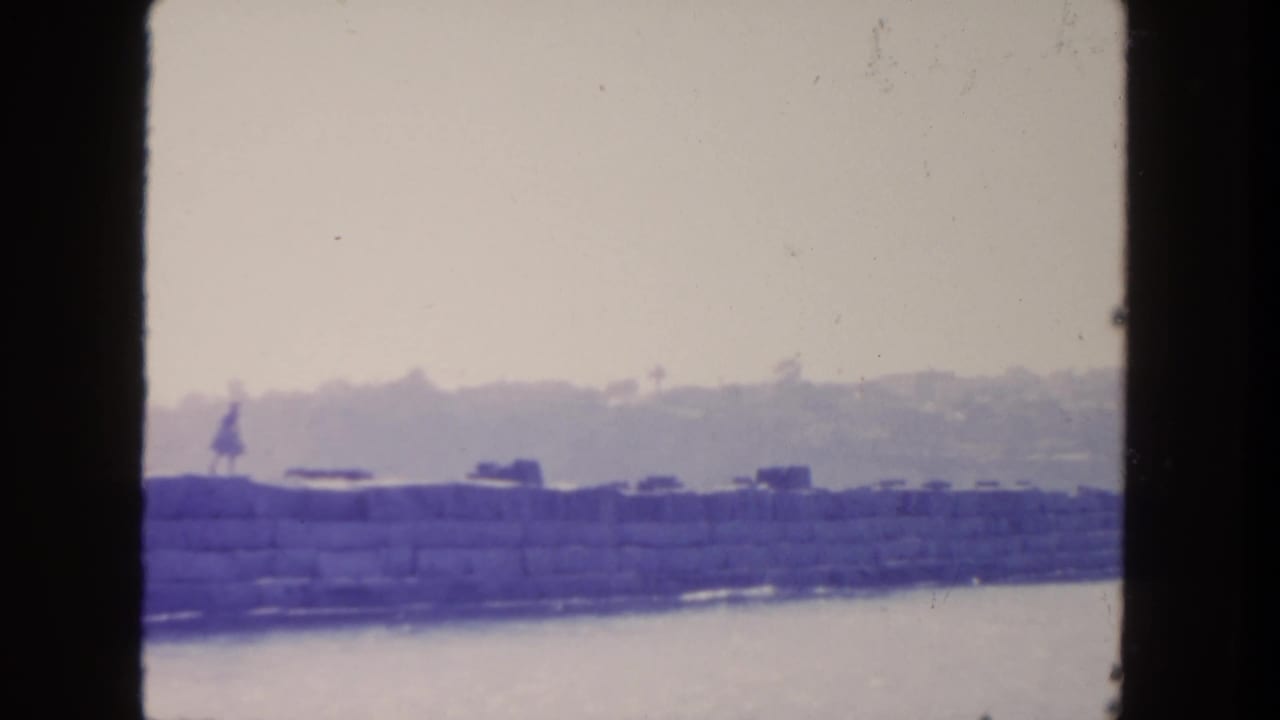 1962 San Diego home-movie frame with a distant woman on a stone seawall, water below, and soft hillside forms in the background.