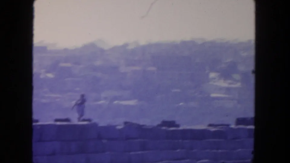 1962 San Diego 8mm frame showing the runner near the lower left, with a stone seawall and misty hillside cityscape behind her.