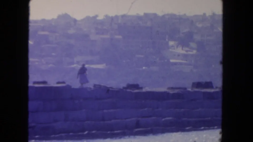 1962 San Diego frame showing a woman in motion on a waterfront seawall, with water and a hazy built hillside in the distance.