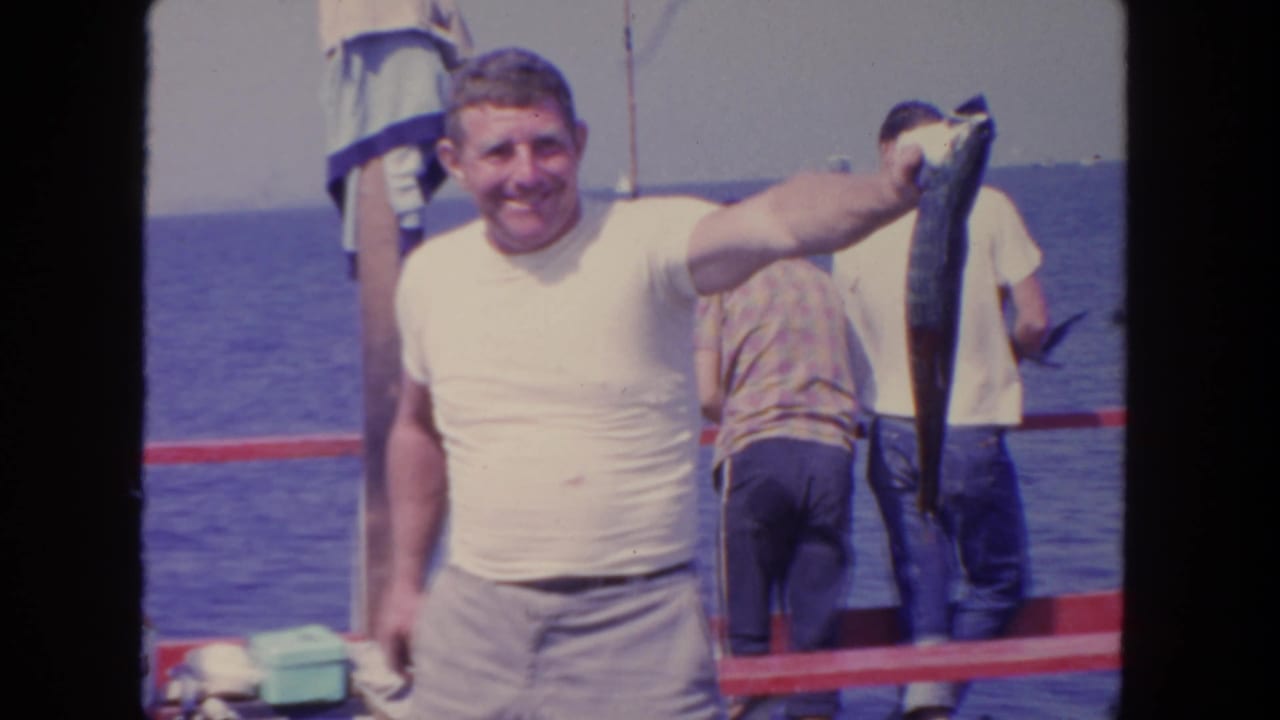 Smiling man in a white T-shirt holds up a large fish on a boat at sea.