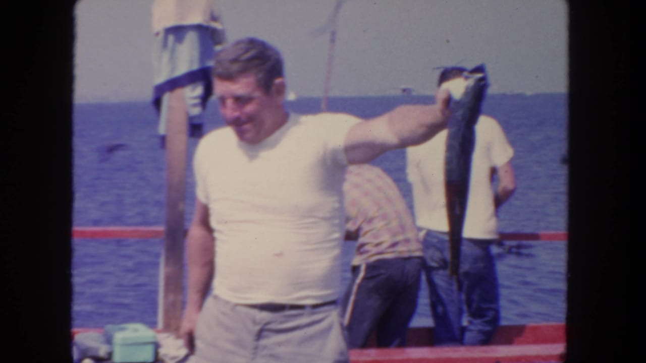 1962 San Diego home-movie frame showing a man in a white T-shirt holding a large fish on a boat with men and sea in the background.