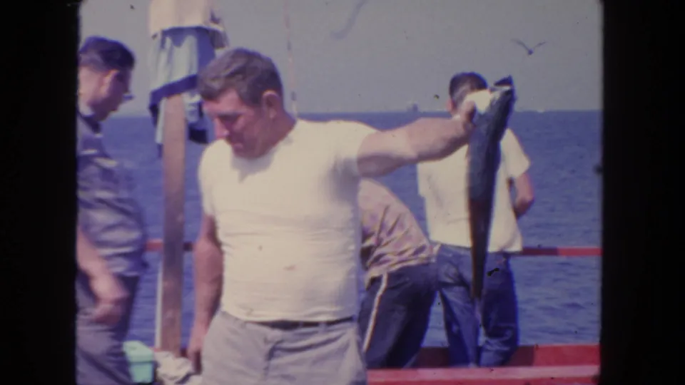 1962 San Diego 8mm frame of a man holding a large fish on deck, with other men, red rail, ocean, and sky visible.