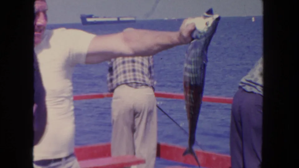 1962 San Diego fishing-boat frame with a large fish held at arm's length, men on deck, ocean water, birds, and a distant ship visible.