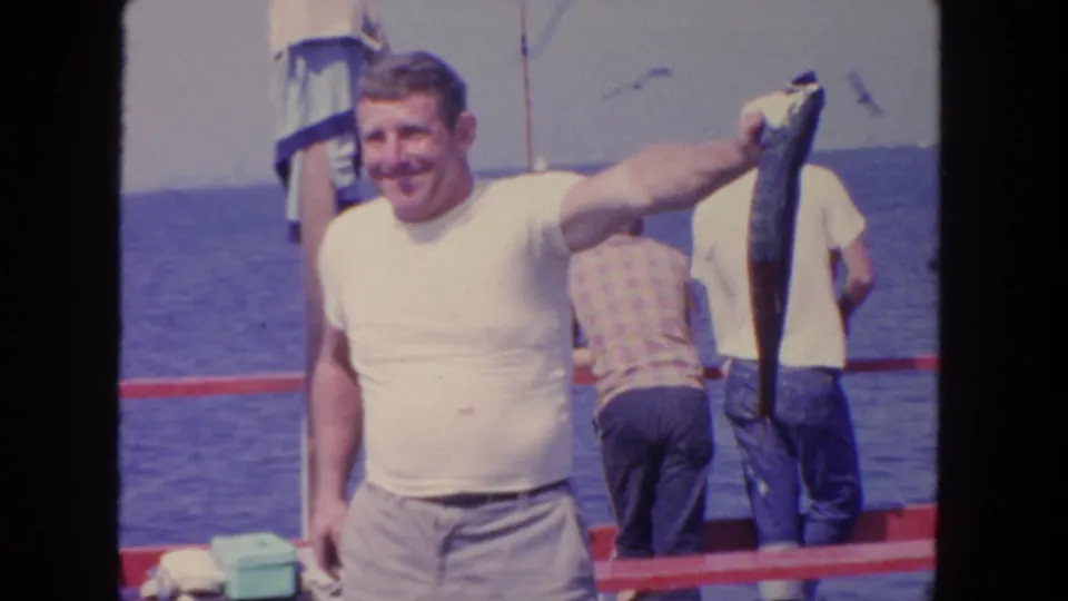 A smiling man in a white shirt displays his fish on a boat, with other anglers and open water visible in San Diego, 1962.