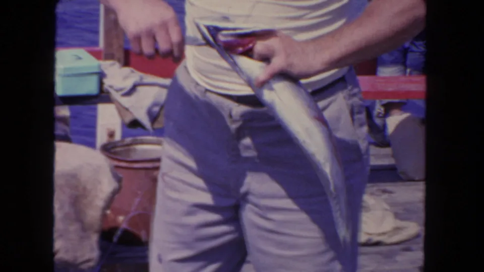 1962 San Diego home-movie frame with a close handheld view of an adult man holding a freshly caught fish on deck.