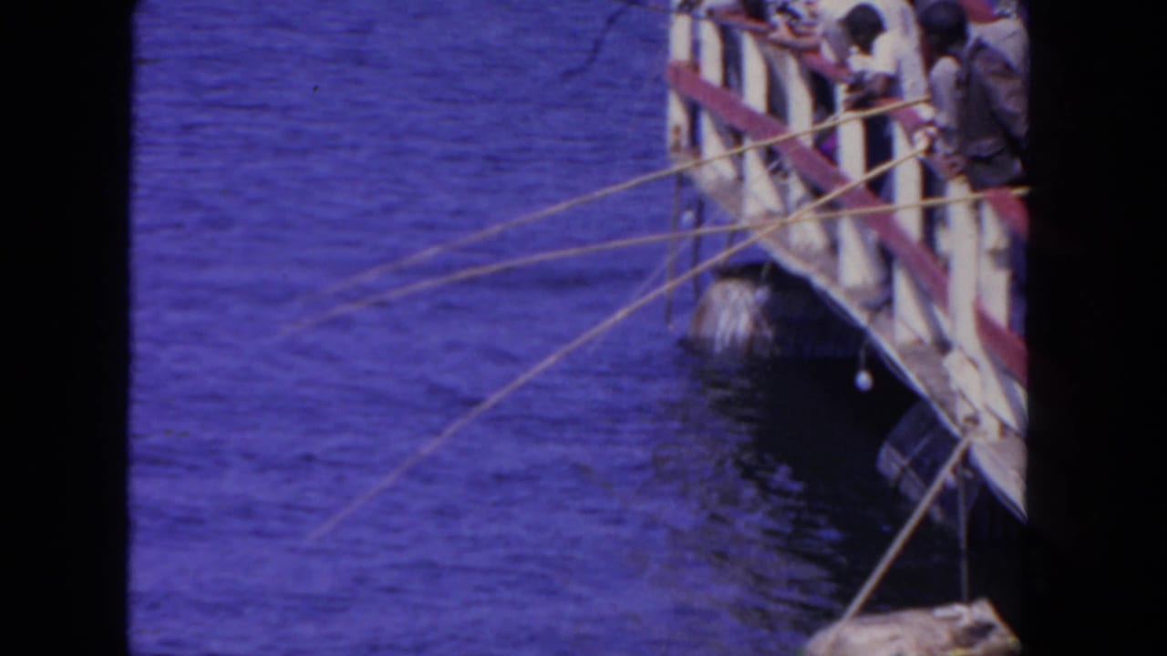 In supported 1962 San Diego home-movie footage, men lean over a wooden platform railing with fishing rods over blue water.