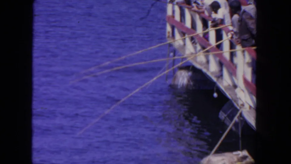 In supported 1962 San Diego home-movie footage, men lean over a wooden platform railing with fishing rods over blue water.