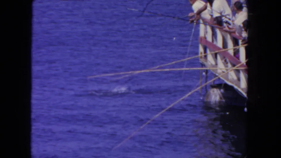 Men in light shirts cluster at a wooden railing, holding fishing rods over blue water with a small splash near the lines.