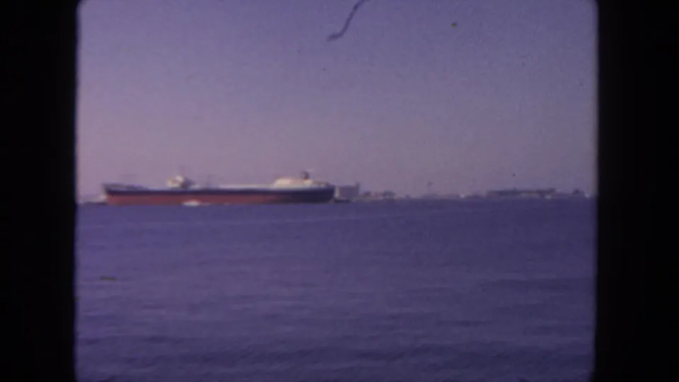 1962 8mm view likely near San Diego showing a large freighter on open water with distant shoreline under a hazy purple sky.