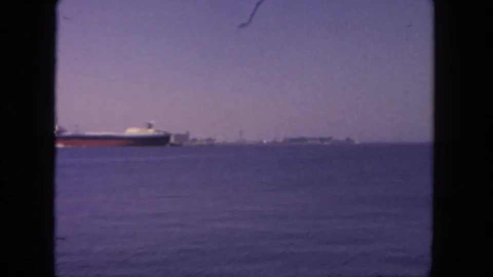 Likely San Diego harbor view from 1962 8mm footage, with a freighter crossing open water and a low skyline on the horizon.