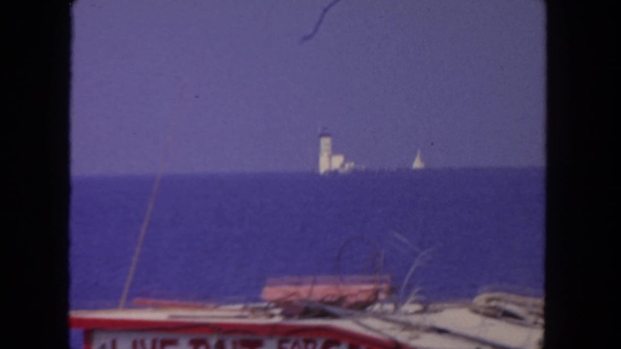 Boat deck foreground with open ocean, distant tower, and small sailboat on the horizon.