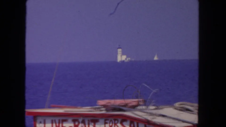 1962 San Diego home-movie view from a fishing boat, with deck hardware, partial signage, ocean, and distant sailboat visible.